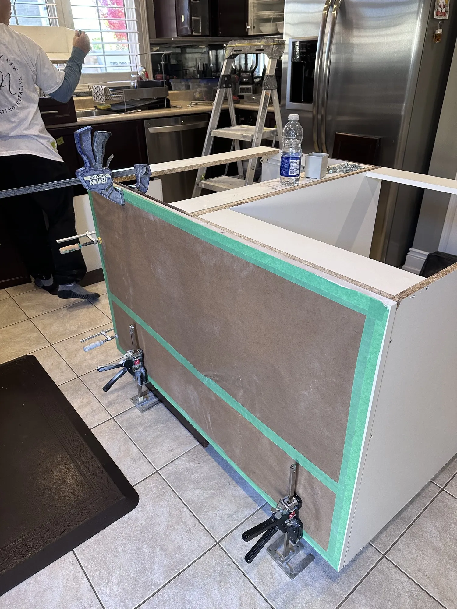 The island end panel clamped and taped in place while the adhesive sets, the new white box visible behind it