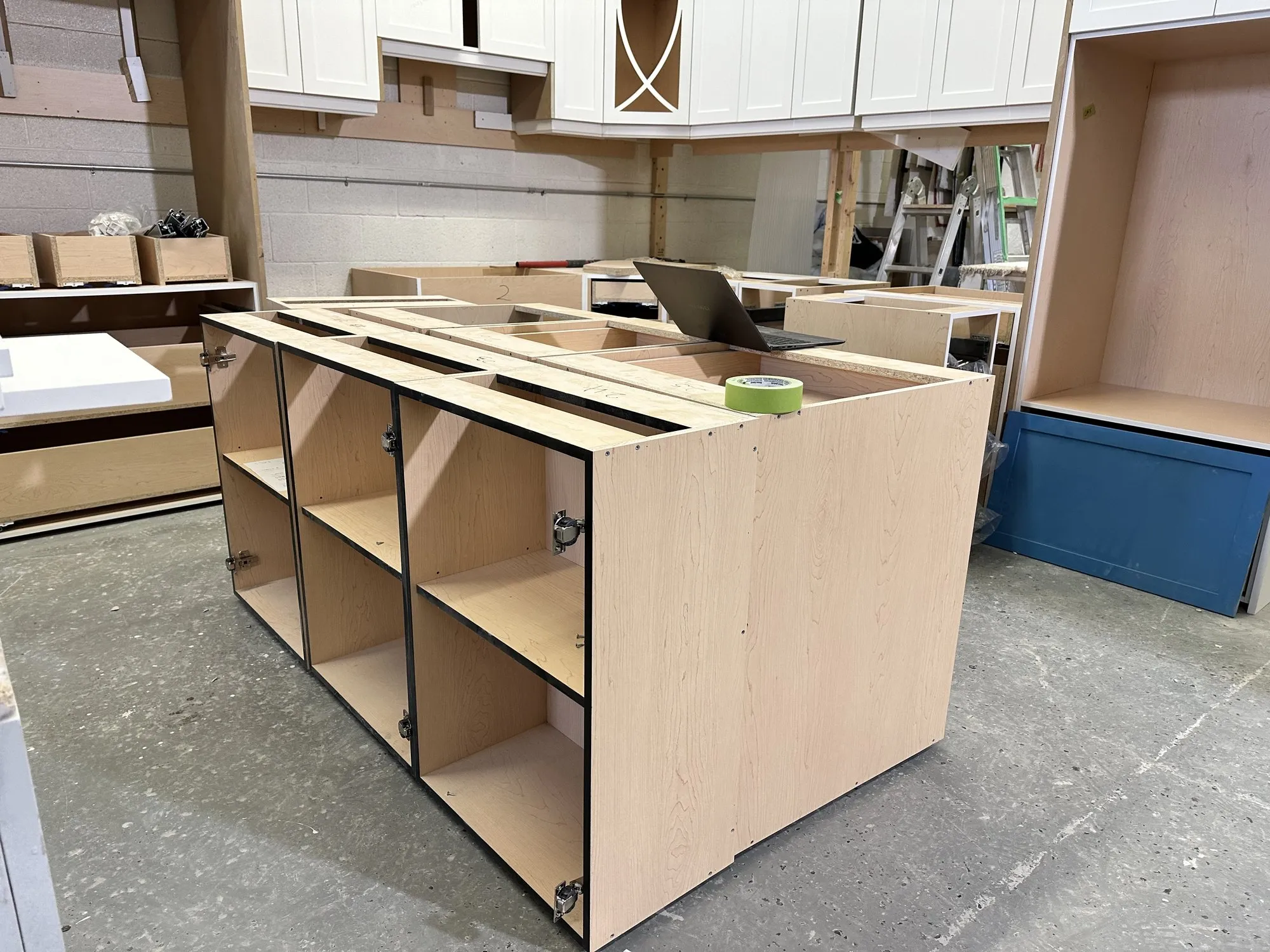 The island box part-built on the shop floor, its plywood compartments open, with the finished blue panels and white mullion-glass doors stacked behind it