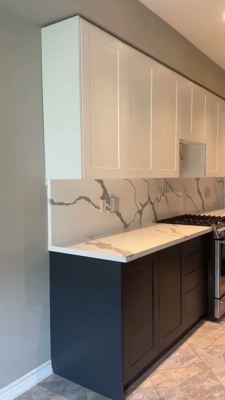 The finished Rouge Woods kitchen — white shaker uppers over charcoal bases, quartz counters with the veining carried up the wall as a slab backsplash, and the original columns kept