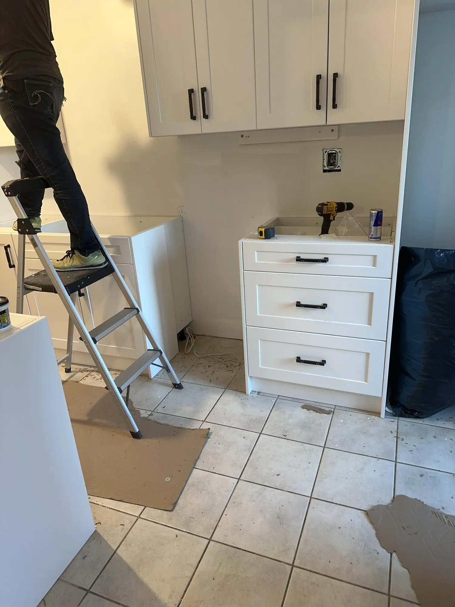 New cabinet boxes being fitted against a bare wall, one being held in place from a stepladder, with the floor cleared